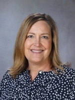 A woman with shoulder-length brown hair, wearing a navy blue blouse with white polka dots, smiles at the camera against a gray background.