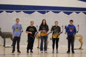 A group of five archers stands together in a gymnasium, each holding a bow, with targets resembling deer in the background.