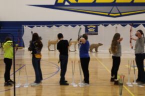 A group of archers stands in a gymnasium, preparing to shoot arrows at targets, with a backdrop featuring animal silhouettes.