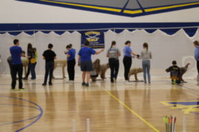 A group of people participating in an archery practice session indoors, with targets set up in the background and a banner hanging on the wall.