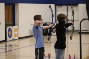 Two young archers are standing side by side in a gym, each drawing their bows and aiming at targets in the background.