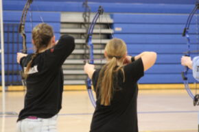 Two individuals are practicing archery indoors, facing away from the camera, with bows drawn and aimed at a target in the distance.