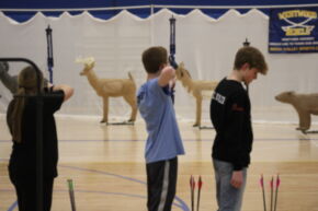Three young archers are positioned in a gymnasium, preparing to shoot arrows at lifelike animal targets in the background.