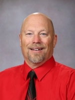 A smiling man with a bald head and a goatee, wearing a red shirt and black tie, poses for a professional headshot against a gray background.