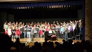 A large group of singers, dressed in various colorful outfits, performs on stage in front of a piano during a choir concert.