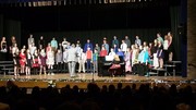 A large group of performers in formal attire is on stage during a choir concert, with a pianist seated at a keyboard and an audience visible in the foreground.