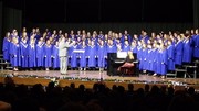 A large choir dressed in blue robes performs on stage, accompanied by a pianist at a grand piano, with an audience visible in the foreground.