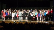 A large choir of singers dressed in colorful outfits performs on stage, accompanied by a pianist, with an audience visible in the foreground.
