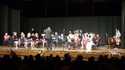 A conductor leads a musical ensemble on stage, with musicians playing various instruments in a performance setting.