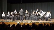 A musical ensemble performs on stage, with a conductor leading a group of musicians dressed in black attire, while an audience watches from below.
