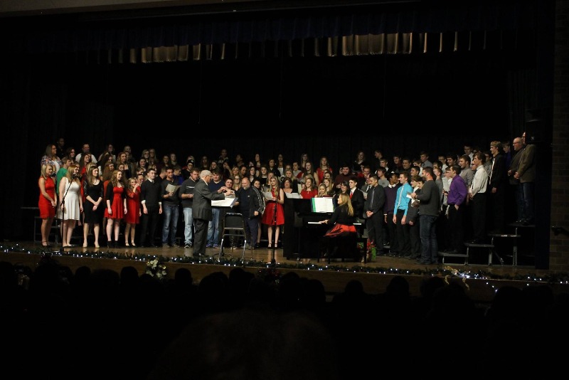 A large choir performs on stage, with a piano and conductor in front, during a concert event.