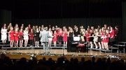 A large group of singers dressed in red and white perform on stage, accompanied by a conductor and a piano, in a concert setting.