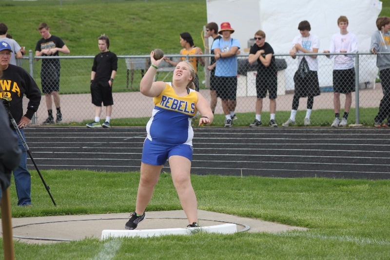 A female athlete in a blue and yellow uniform is preparing to throw a shot put during a track and field event, with spectators in the background.