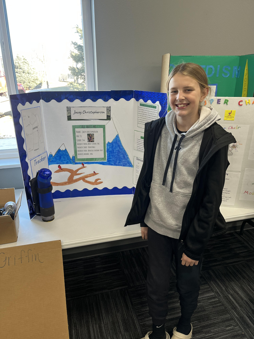 A young girl stands next to a colorful project display board featuring mountains and a tree, with various written sections and a QR code, in a well-lit room.