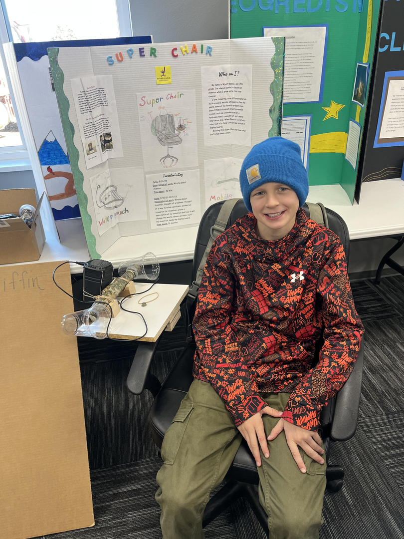 A young boy wearing a blue beanie and a red and black patterned jacket sits in a chair in front of a science fair project display board, which includes various drawings and text.