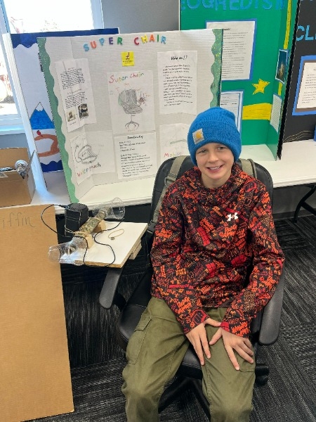 A young boy wearing a blue beanie and a patterned jacket sits in a chair in front of a science project display board, which includes various drawings and text about a project titled 'Super Chair.'