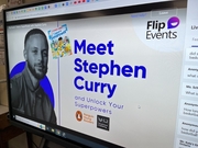 A digital display featuring a promotional event titled 'Meet Stephen Curry' with a black and white image of Stephen Curry, accompanied by colorful graphics and text inviting viewers to unlock their superpowers.