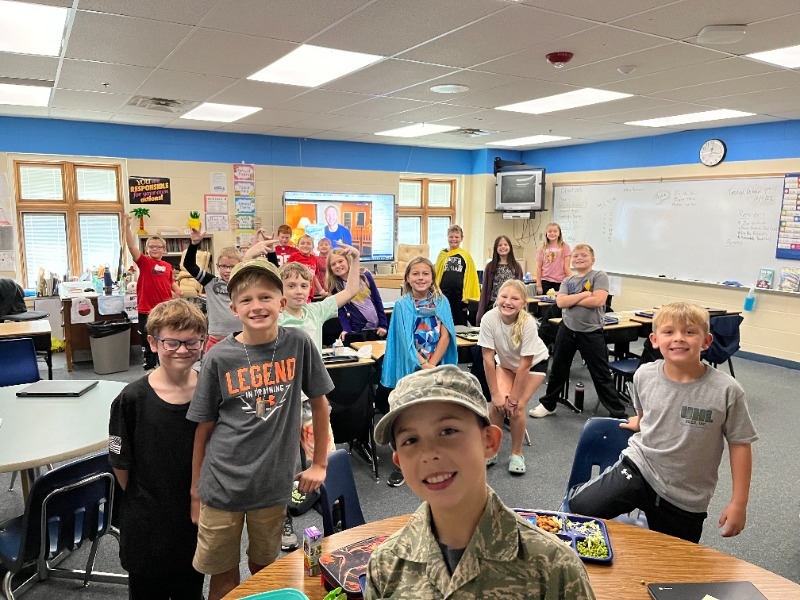 A group of students smiling and posing for a photo in a classroom, with desks and educational materials visible in the background.