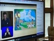 A digital presentation featuring a colorful illustrated scene with children and dogs in a park, alongside video feeds of two individuals, one signing and the other speaking, displayed on a large screen.
