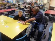 A child and an older man in a wheelchair are engaged in a reading activity at a brightly colored table in a library, surrounded by bookshelves filled with books.