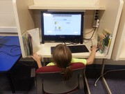A child with long hair is seated at a computer desk, focused on a monitor displaying educational content, with books organized on either side of the desk.