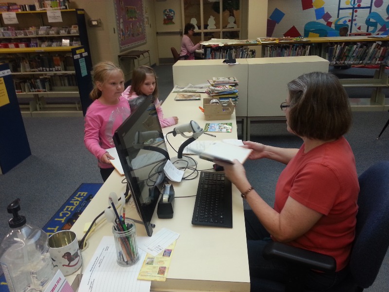 A librarian assists two young girls at a library checkout desk, with books and a computer visible in the background.