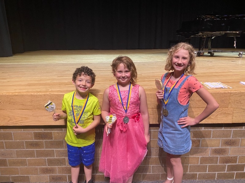 Three children stand on a stage, each holding a trophy and wearing medals, with a piano visible in the background.