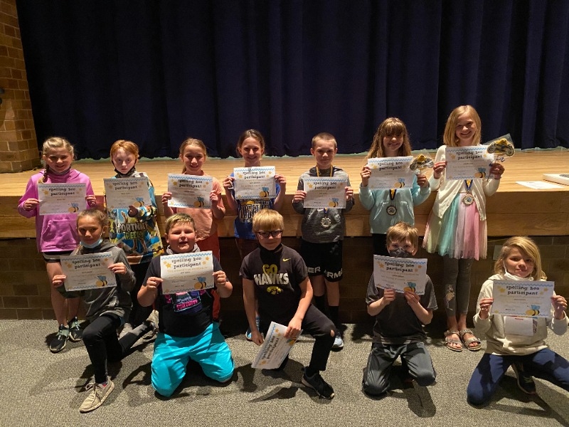 A group of children holding certificates stands in front of a stage with a blue curtain, smiling for the camera.