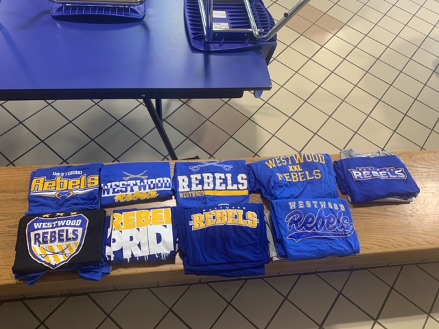 A collection of blue and yellow t-shirts displaying various designs and text related to 'Westwood Rebels' arranged neatly on a wooden bench in a cafeteria setting.