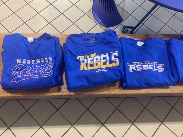 A display of folded blue t-shirts featuring the text 'Westfield Rebels' in various styles, arranged neatly on a wooden shelf.