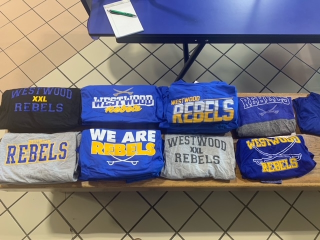 A display of folded t-shirts in various colors, featuring the words 'REBEL', 'WE ARE REBELS', and 'WESTWOOD REBELS' in different styles and sizes, arranged neatly on a table.