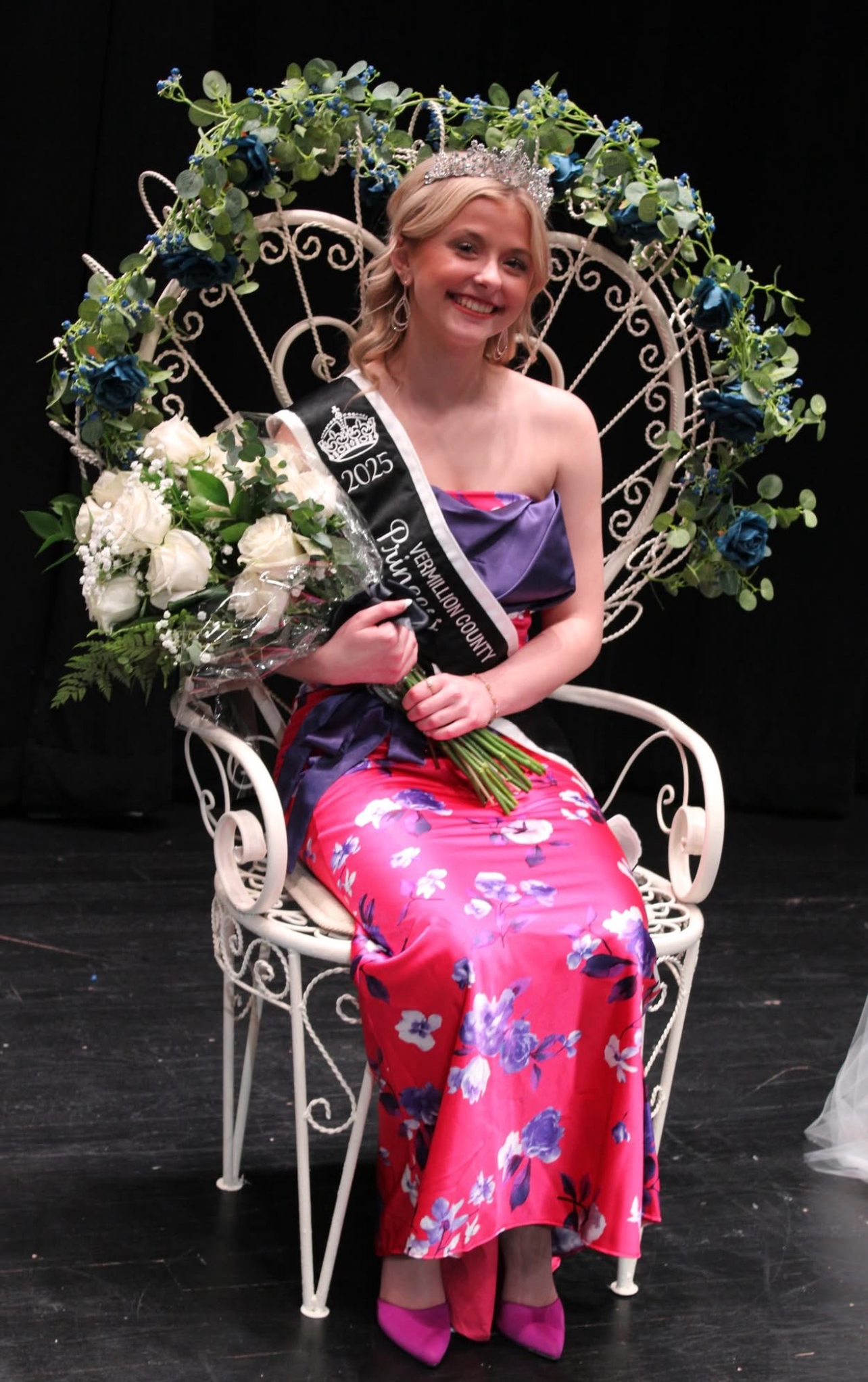 girl sitting in a chair wearing a sash and crown and holding a bouquet of flowers