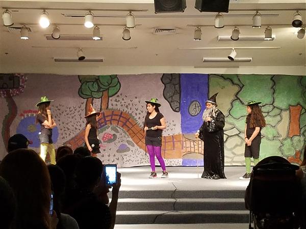 A group of children dressed in costumes perform on stage in front of a colorful backdrop depicting a whimsical landscape.