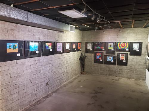 An art gallery featuring a series of framed artworks displayed on a textured wall, with a small plant in the corner and overhead lighting illuminating the pieces.
