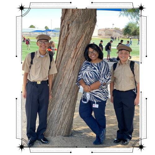 Two students in military-style uniforms stand beside a woman in a striped shirt, all posing near a tree on a school campus with other students in the background.