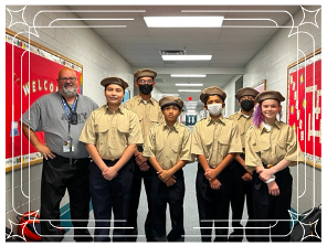 A group of six students wearing beige uniforms and hats stands in a school hallway alongside an adult, all smiling for the camera.