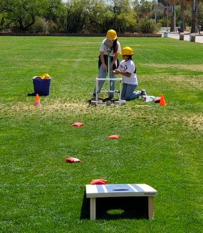 Two individuals wearing yellow helmets are engaged in an outdoor activity on a grassy field, with one using a tool while the other assists, and a cornhole game setup in the foreground.