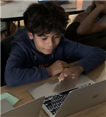A young boy with curly hair is focused on a laptop, resting his chin on his hands, while other students are visible in the background.