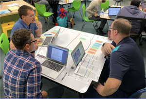 Three individuals are engaged in a discussion around a table with laptops and documents, in a collaborative workspace filled with green chairs.