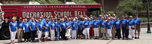 A large group of people wearing blue shirts stands in front of a red bus with the words 'Operation School Bell' displayed prominently.