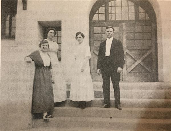 A historical black-and-white photograph of four individuals standing on the steps of a building, with two women in long dresses and two men in formal attire.