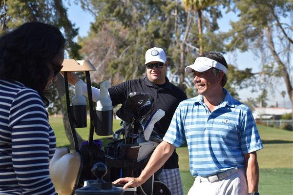 Three individuals are engaged in conversation on a golf course, with one person wearing a blue striped shirt and a visor, while another is in a black shirt and a cap, and a golf bag is visible in the background.