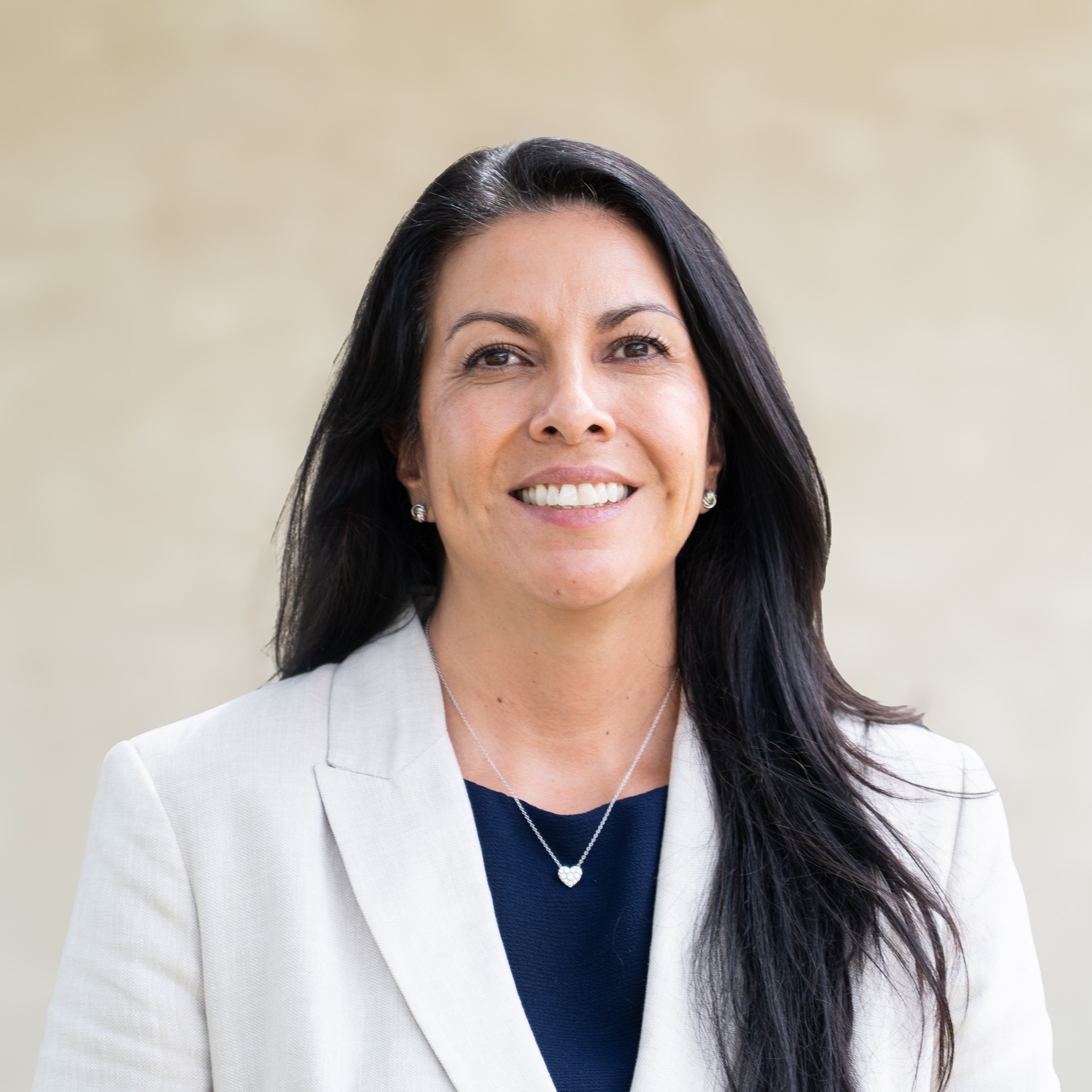 A woman with long black hair smiles, wearing a white jacket and blue top. A light-colored wall in the background.
