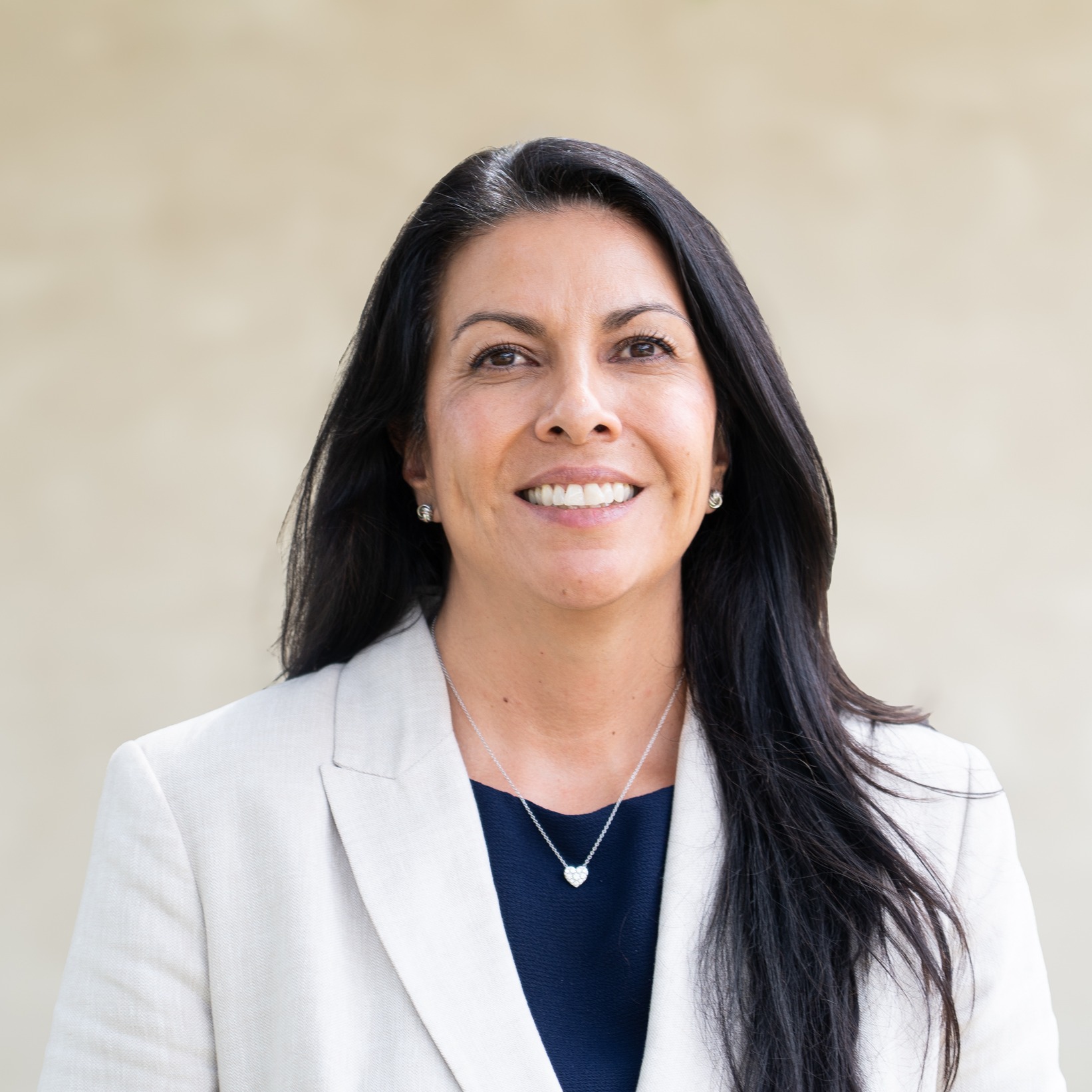 Woman in white blazer, long black hair, and a necklace smiles. Light beige background.