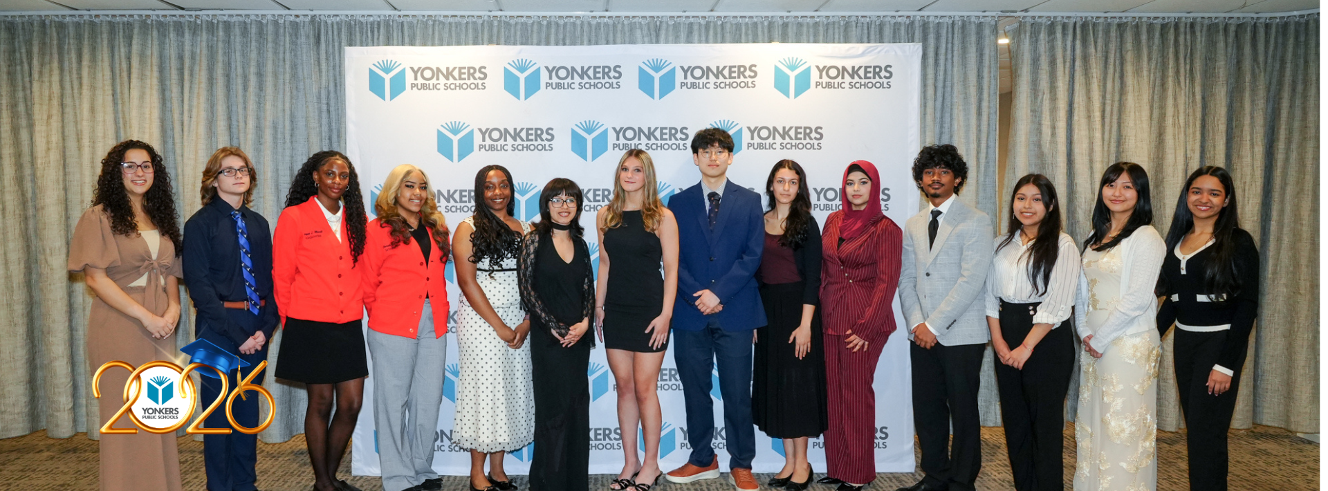 Fourteen of the 16 valedictorians and salutatorians from Yonkers Public Schools—stand in a line, dressed in formal attire, posing in front of a Yonkers Public Schools backdrop at the 2026 recognition event.