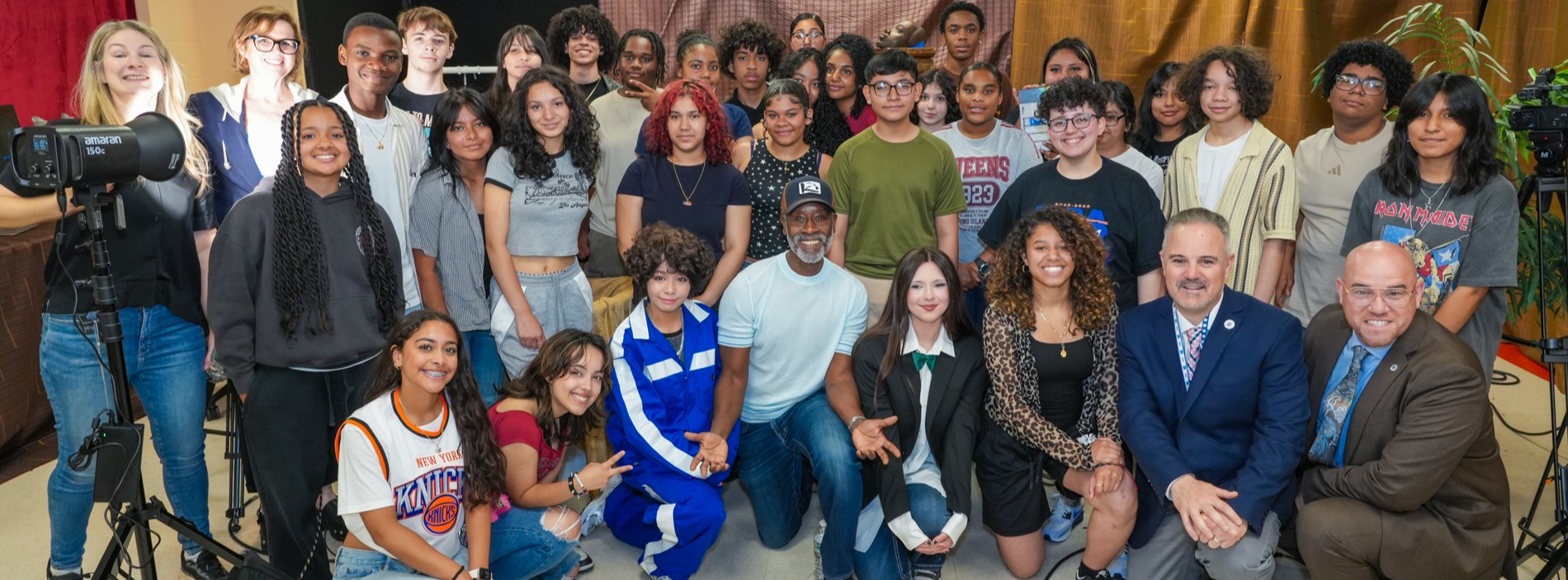 Actor Don Cheadle, wearing a white T-shirt and baseball cap, kneels in the front row surrounded by students, educators, and administrators from the Robert Halmi Sr. Academy of Film and Television during a group photo