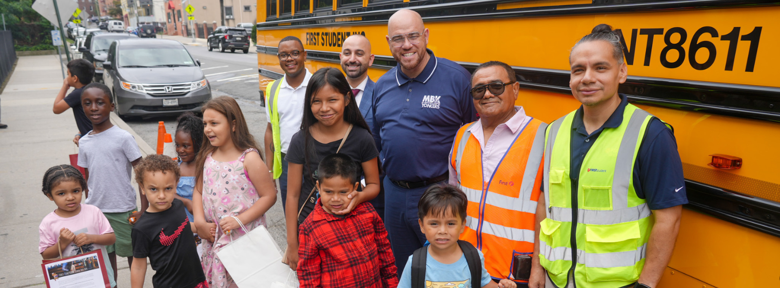 Children preparing to ride the school bus for the first time gather with their experienced older siblings beside a yellow school bus. The superintendent, school principal, bus driver and transportation safety personnel join them for the first-time rider event.