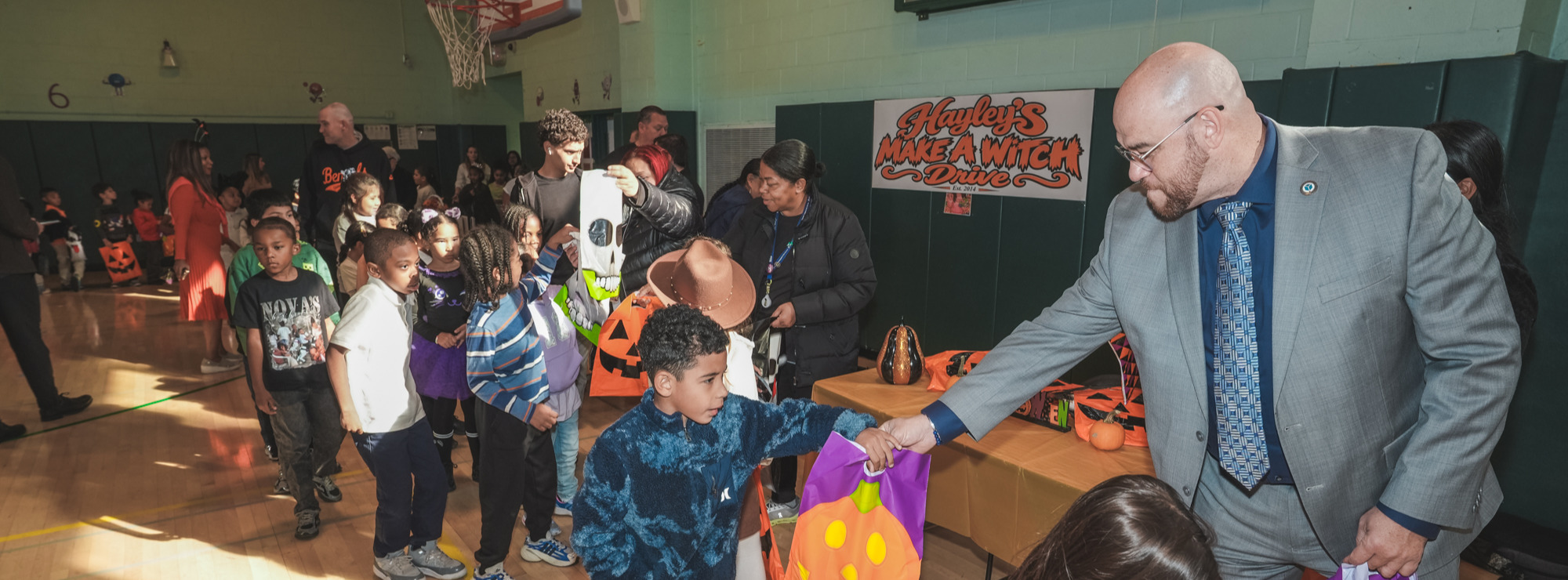 Superintendent Soler, dressed in a grey suit, hands a Halloween treat bag decorated with a jack-o'-lantern to a young boy in a school gymnasium. Other children and adults gather nearby with Halloween bags and costumes beneath a banner reading “Hayley’s Make A Witch Drive.”
