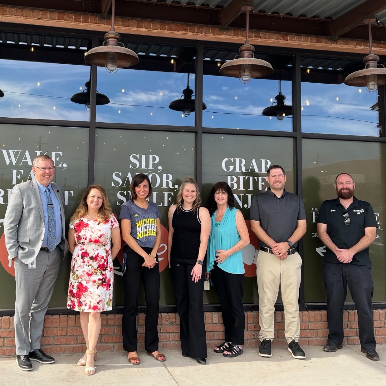 Seven people pose in front of a building with signs that read "WAVE," "SIP," and "GRAB" in the windows.