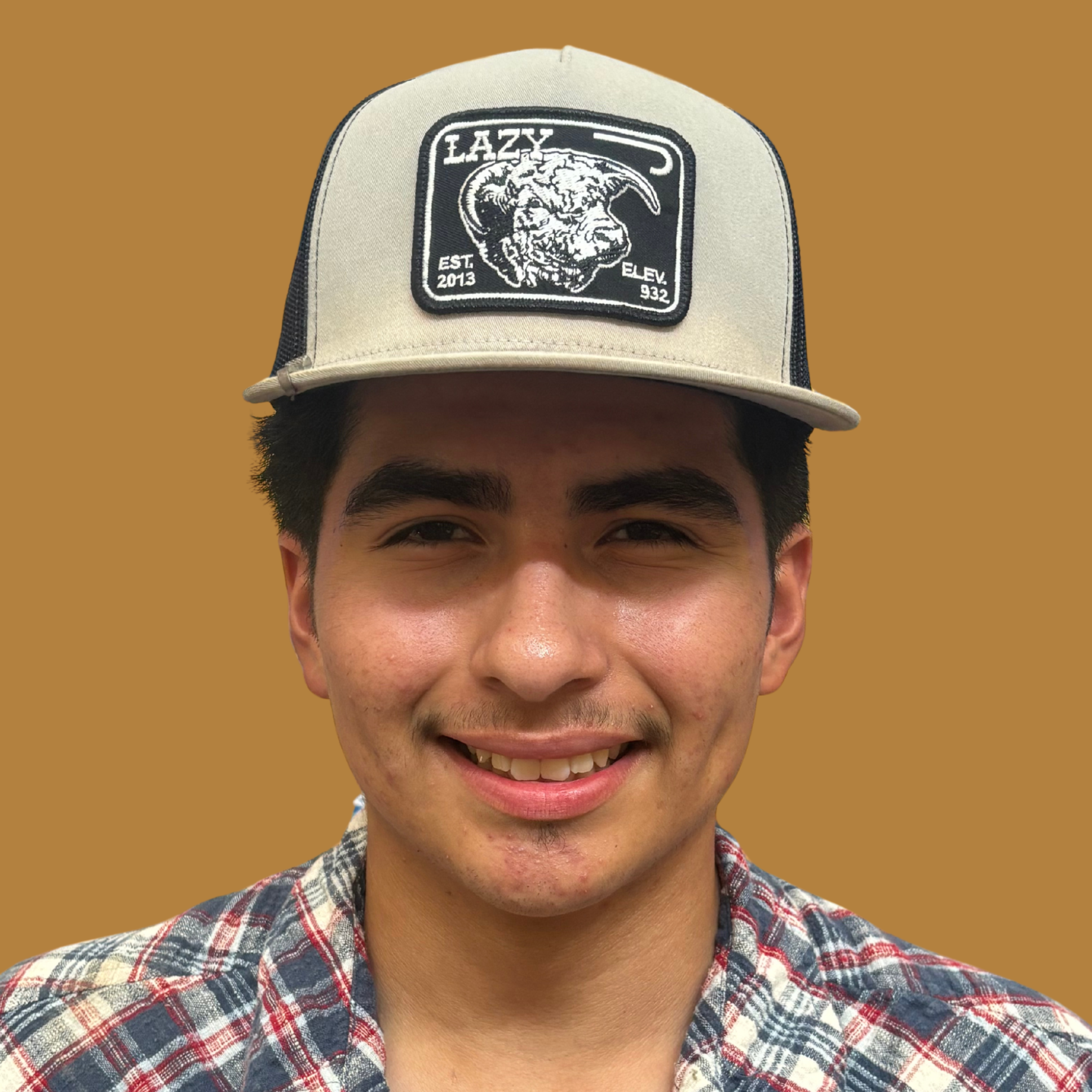 A person in a plaid shirt and cap with a logo smiles at the camera. Brown background.